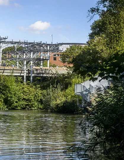 Sjö omgiven av gröna träd, metallkonstruktioner i bakgrunden. Foto.